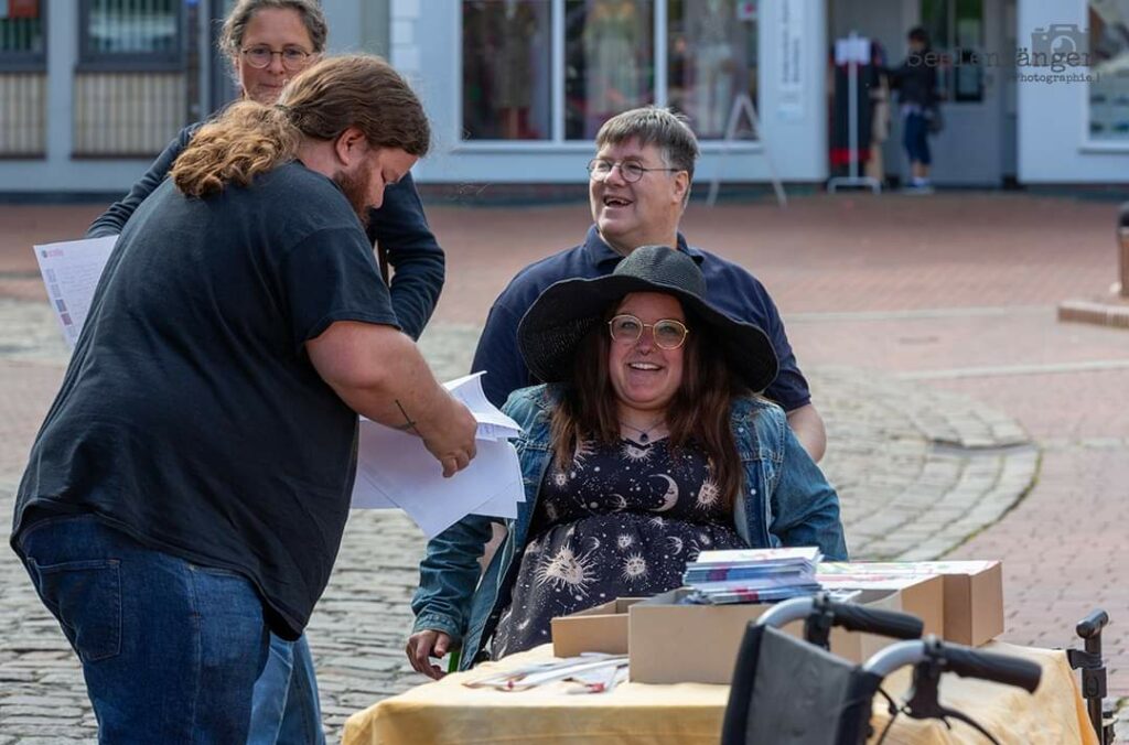 Zwei Rollstuhlfahrende Personen und zwei laufende Personen auf dem Bild. Im Vordergrund ist ein Tisch mit Materialien zu sehen.