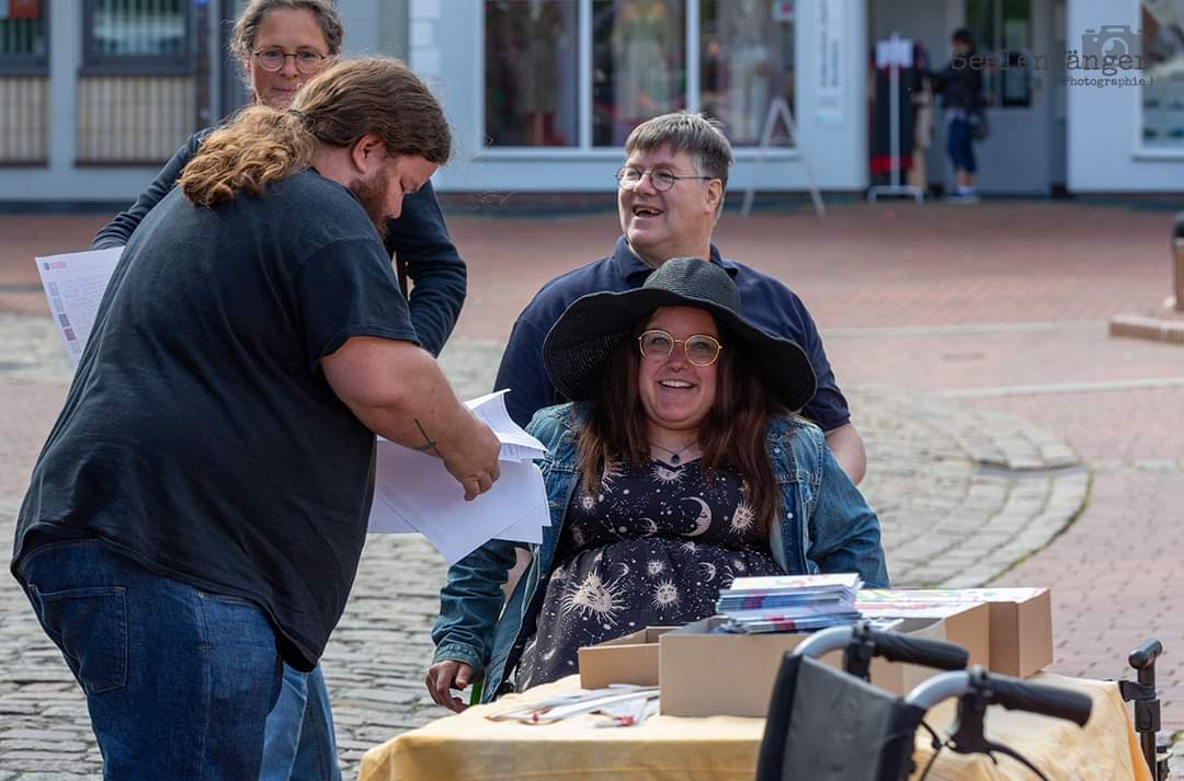 Zwei Rollstuhlfahrende Personen und zwei laufende Personen auf dem Bild. Im Vordergrund ist ein Tisch mit Materialien zu sehen.